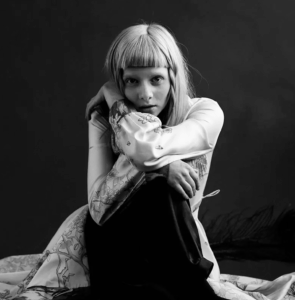 Photo of the norwegian musician Aurora. She is sitting on the ground and wearing a dress.